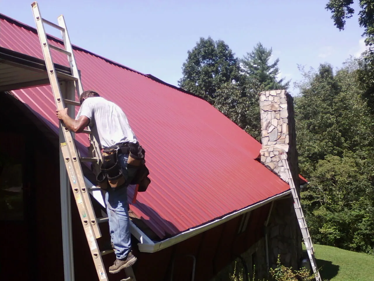 Roofer on a ladder beside a red metal roof and stone chimney — Soffit & Fascia Repair by Roofing Brothers in Greensburg